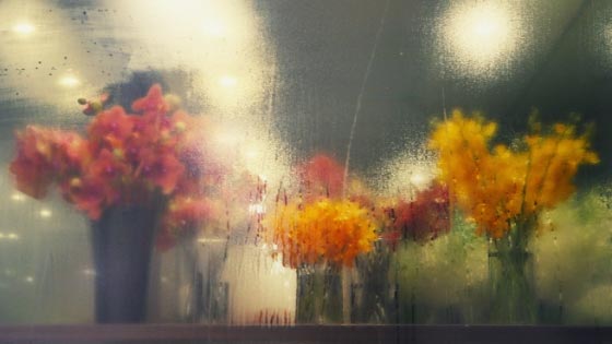 A moist glass wall of a flower shop and inside of the shop are a bunch of colorful flowers placed inside the vases